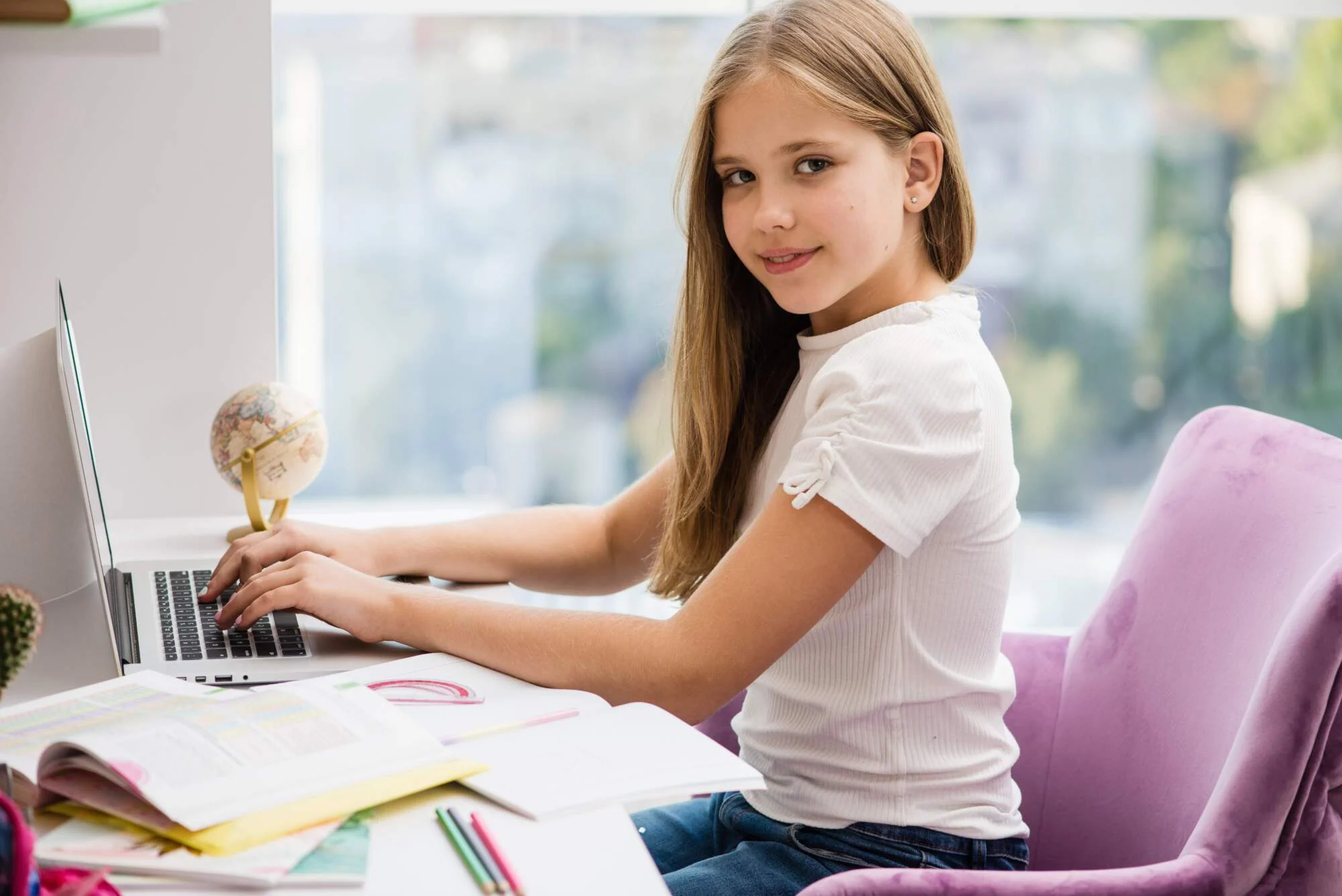 Young girl with long blonde hair sitting at a desk with a laptop, globe, and study materials, looking at the camera with a slight smile