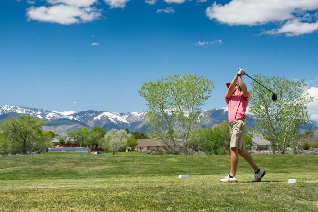 A golfer in a pink shirt follows through on a drive from the tee, with snow-capped mountains beyond
